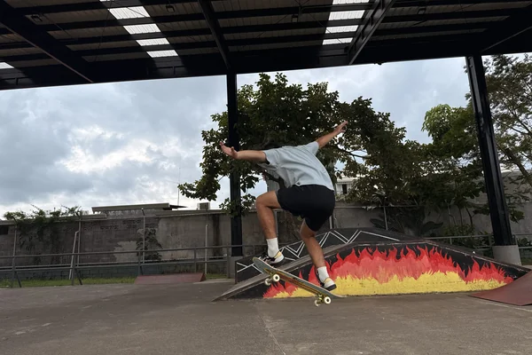 A young person is performing a skateboard trick in a park setting. They are captured mid-air, having just jumped off a ramp. The skateboarder is wearing a light blue shirt and black shorts, with their legs bent and arms extended for balance. Below them, there’s a colorful ramp designed with a fiery gradient of red, orange, and yellow.