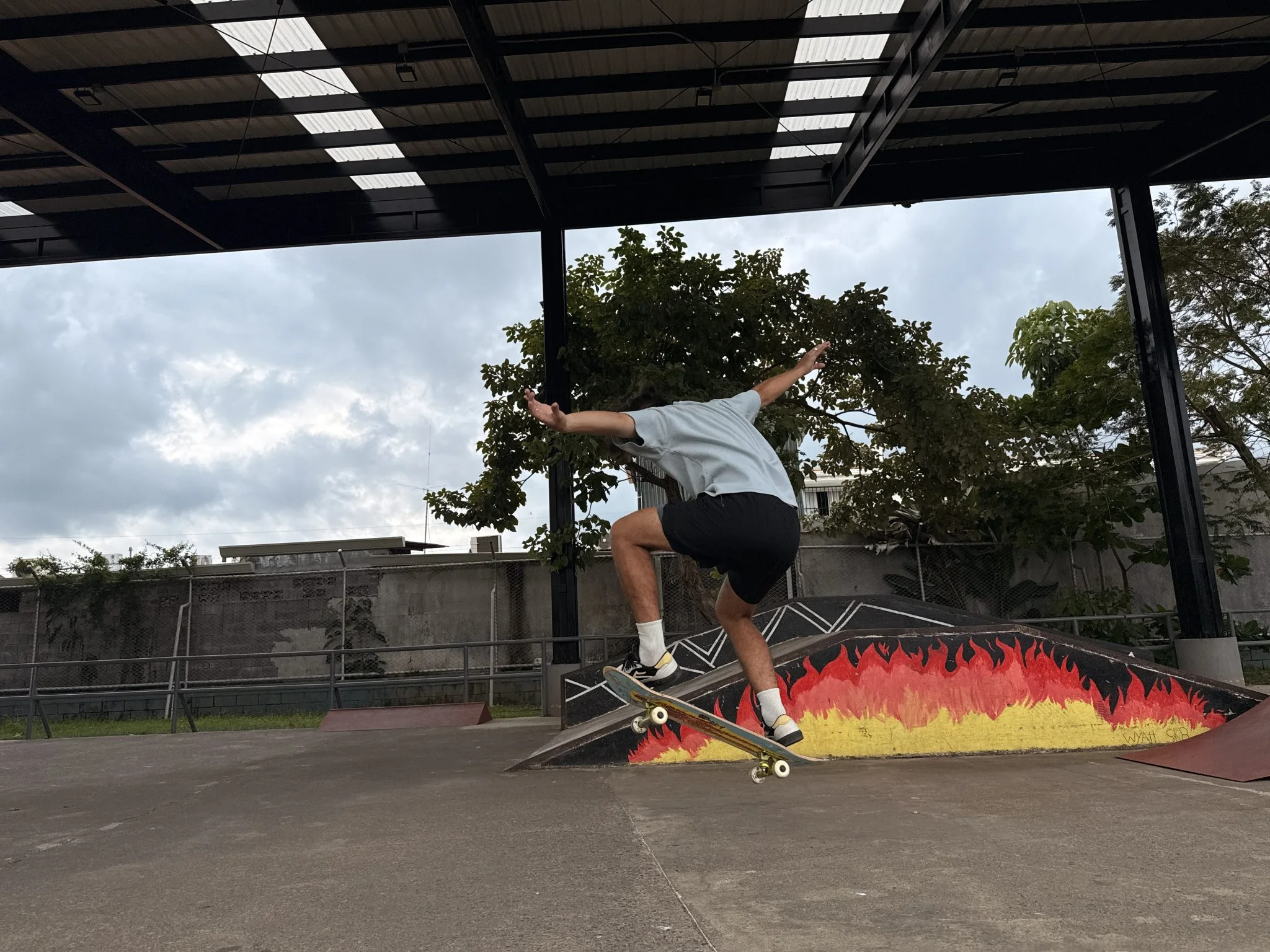 A young person is performing a skateboard trick in a park setting. They are captured mid-air, having just jumped off a ramp. The skateboarder is wearing a light blue shirt and black shorts, with their legs bent and arms extended for balance. Below them, there’s a colorful ramp designed with a fiery gradient of red, orange, and yellow.